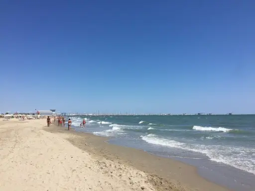 Spiaggia affollata a Ravenna, con gente che passeggia sulla sabbia e nel mare. All'orizzonte, barche e un cielo azzurro intenso.