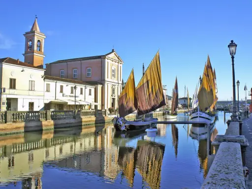 Traditional sailboats with colorful sails line a canal in Cesenatico, Italy, with buildings and a church reflected in the water.