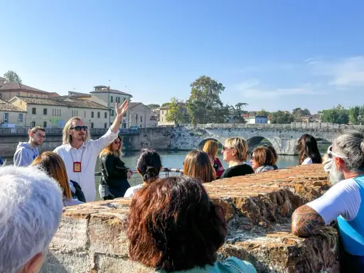 Guida turistica con gruppo a Rimini, che ascolta storie sui **eventi rimini** e la storia locale, con un ponte antico sullo sfondo.