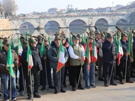 Uomini con cappelli alpini e bandiere italiane partecipano all'adunata alpini a Rimini, con un ponte storico sullo sfondo.