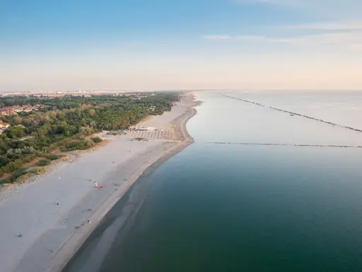 Vasta spiaggia dorata e mare calmo, con file di ombrelloni che delineano un lido di classe. All'orizzonte, la costa punteggiata di verde e costruzioni.