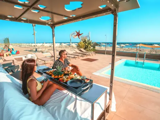 Coppia rilassata su lettini con vassoio di frutta e caffè, con piscina e spiaggia di Rimini sullo sfondo.