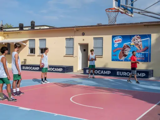 Ragazzi in tenuta sportiva giocano a basket in un camp estivo a Cesenatico, con un cartellone pubblicitario di gelati Motta sullo sfondo.