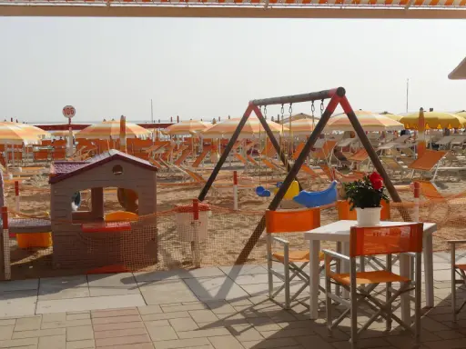 Area giochi e lettini sulla spiaggia 106 di Riccione, con ombrelloni arancioni e bianchi, altalena e casetta per bambini.