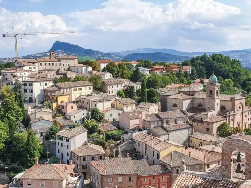 Panoramica di un borgo collinare con tetti rossi e una chiesa con campanile, incorniciato dal verde e dalle montagne. Un paesaggio che evoca la bellezza di Rimini e dintorni.