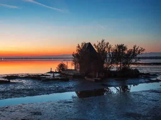 Tramonto sulla spiaggia di Marano, con una piccola capanna e un'imbarcazione riflessi sull'acqua calma.