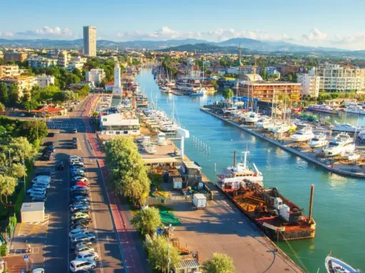 Vista aerea del porto di Rimini con barche ormeggiate, edifici e montagne sullo sfondo.