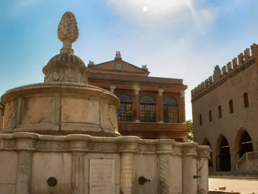 Fontana storica nel centro di Rimini, con edifici antichi sullo sfondo e cielo azzurro.