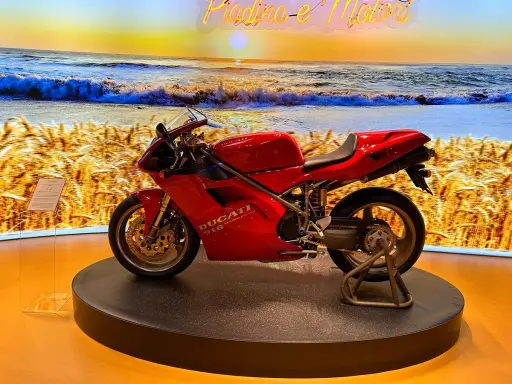 Ducati rossa su un palco, con sfondo di un campo di grano e il mare al tramonto. Un connubio perfetto tra la piada e il mare.