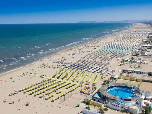 La riviera di Rimini in pieno sole, con un mare azzurro e una spiaggia affollatissima di ombrelloni colorati e bagnanti.
