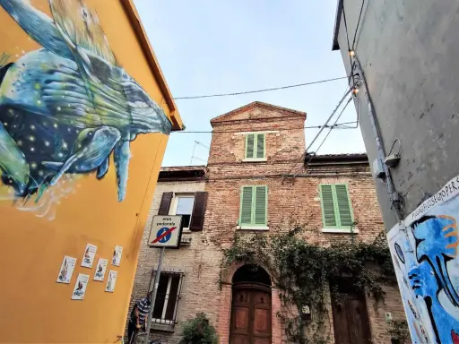 Un murale di balena colorata decora un edificio giallo a San Giuliano a Mare. Vicino, un edificio in mattoni con persiane verdi e un cartello di area pedonale.