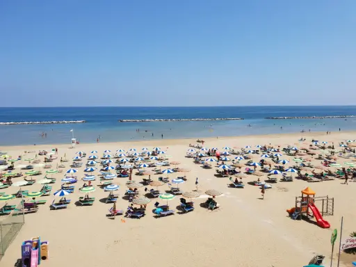Spiaggia libera Viserba affollata di ombrelloni e lettini sotto un cielo azzurro. Bambini giocano nell'area attrezzata.