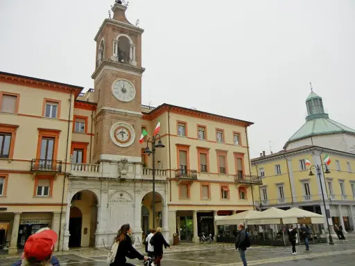La piazza Rimini, con il suo imponente campanile e gli edifici storici, accoglie visitatori e residenti sotto un cielo nuvoloso.