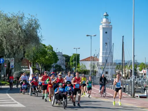 Atleti corrono lungo il percorso della Rimini Marathon, con il faro bianco e il porto sullo sfondo.