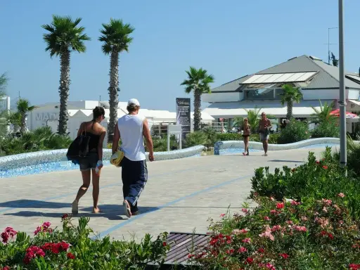Coppia passeggia sul lungomare del parco mare nord Misano, tra palme e fiori.