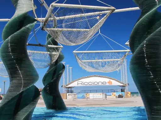 Sculture di vetro e reti sospese, con la scritta "Riccione" in lontananza. Un'idea di cosa vedere a Riccione, tra arte e mare.