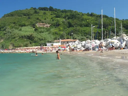 Spiaggia affollata con gente che nuota e prende il sole. Un promontorio verde domina la scena. Meglio Riccione o Gabicce Mare? Difficile scegliere!