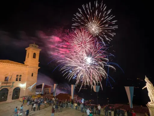 Spettacolo pirotecnico illumina il cielo notturno per la festa di San Marino. Folla ammira i fuochi d'artificio da una piazza con un edificio storico.