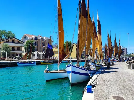 Barche a vela ormeggiate in un porto con case colorate sullo sfondo. Un dilemma: meglio Bellaria o Igea Marina?