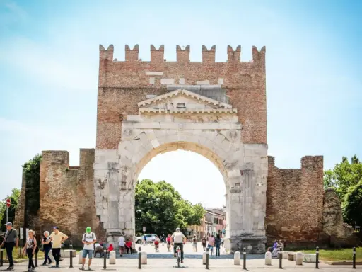 L'imponente arco d'augusto rimini, con le sue antiche pietre e la torre in mattoni, fa da sfondo a una vivace piazza con persone che passeggiano e ciclisti.