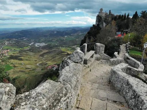 Sentiero in pietra conduce al **San Marino castle**, con vista panoramica sulla valle verdeggiante e colline ondulate sotto un cielo nuvoloso.
