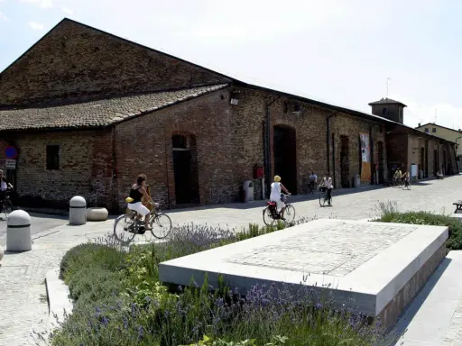Passeggiata in bicicletta davanti ai Magazzini del Sale di Cervia, con persone che pedalano su un lungomare acciottolato, tra aiuole fiorite e antichi edifici in mattoni.
