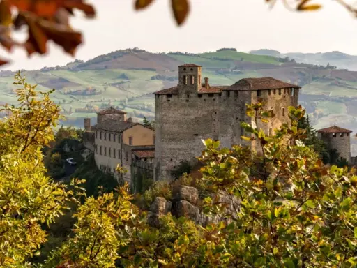 Castello medievale su una collina, circondato da alberi con foglie autunnali dai colori caldi. Il paesaggio collinare sullo sfondo evoca un senso di pace in questo mare autunno.