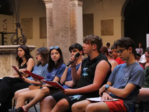 Giovani ascoltano un discorso, alcuni con fogli rossi in mano, durante un evento culturale a Rimini. Un ragazzo tiene un microfono.