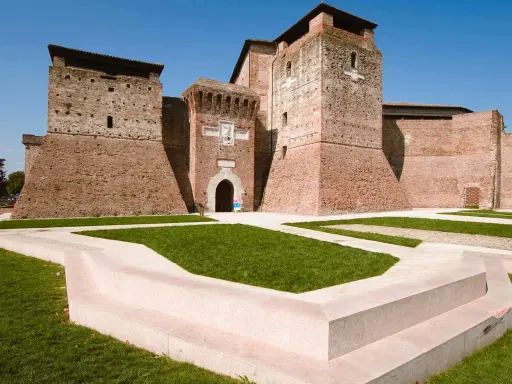 Imponente Castel Sismondo, con le sue torri in mattoni rossi e il cortile verdeggiante, si erge maestoso sotto un cielo azzurro.