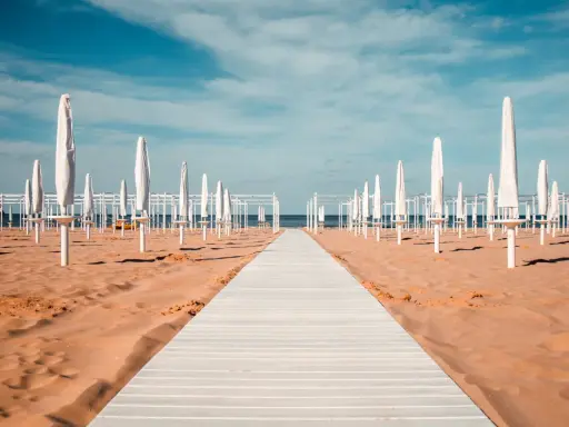Passerella bianca su sabbia dorata, file di ombrelloni chiusi e strutture bianche sotto un cielo azzurro. Un'immagine serena del mare in Emilia-Romagna.