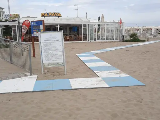 Percorso a strisce bianche e blu sulla sabbia del piazzale San Martino a Riccione, conduce al bar Papaya e alla spiaggia attrezzata.