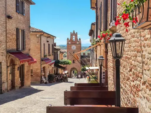 Un vicolo acciottolato a Gradara, con edifici in mattoni, fiori rossi e un campanile con orologio. Perfetto per scoprire cosa vedere.