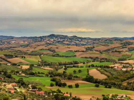 Dolci colline verdi e marroni della Valconca, Rimini, punteggiate da case e alberi, sotto un cielo nuvoloso.