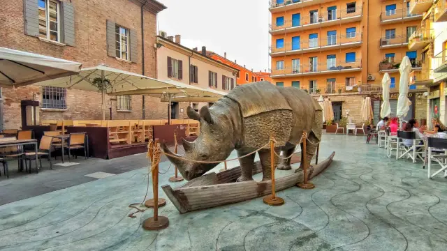 Statua di rinoceronte in una barca, in una piazza di Rimini, dove è nato Fellini. Tavolini all'aperto e palazzi colorati sullo sfondo.
