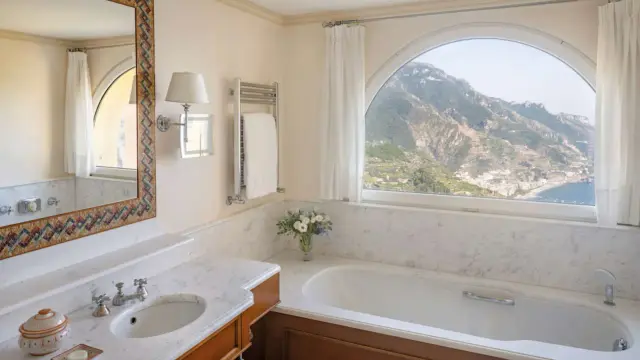 Un bagno di lusso con vasca, lavandino e specchio decorato, che offre una vista mozzafiato sulle montagne e sul mare. Tra i bagni più belli ed eleganti.