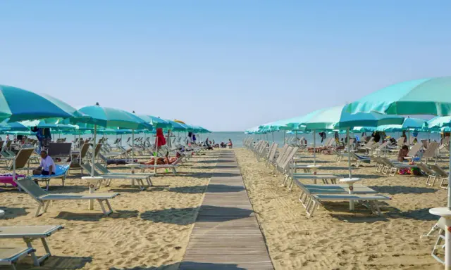 Una passerella di legno conduce alla spiaggia di San Giuliano Mare, fiancheggiata da file di lettini e ombrelloni azzurri sotto un cielo limpido.