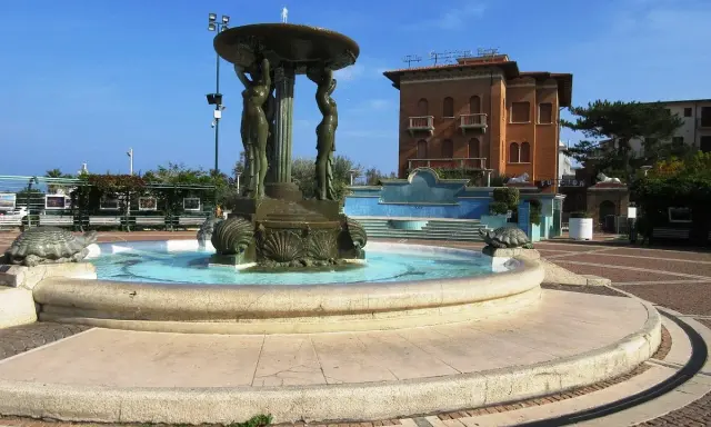 Fontana con statue di donne e tartarughe in piazza Primo Maggio Cattolica, con un edificio in mattoni sullo sfondo.