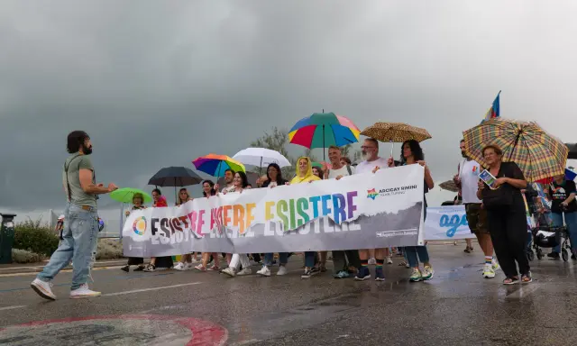 Persone con ombrelli colorati sfilano a Rimini Pride, tenendo uno striscione arcobaleno con scritto "Esistere Mere Esistere".