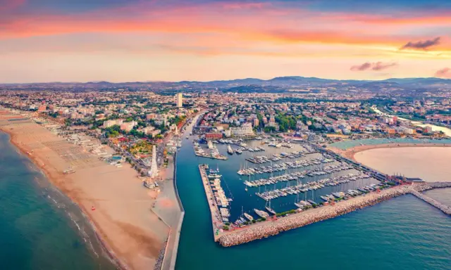 Tramonto sul lungomare di Rimini, con la spiaggia dorata, il porto turistico pieno di barche e la città che si estende fino alle colline.