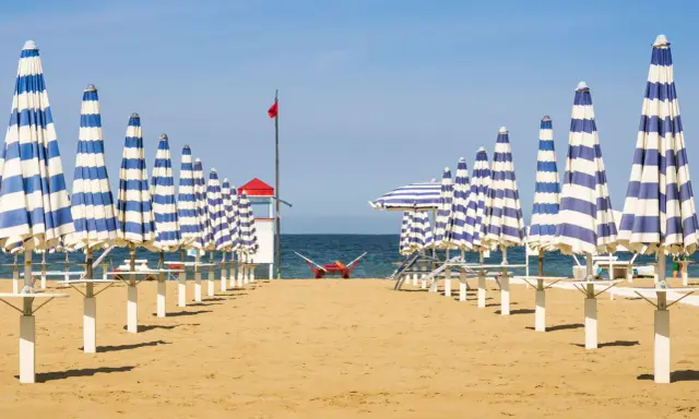 Filetto di ombrelloni a righe blu e bianche sulla spiaggia di Rivabella di Rimini, con mare calmo e cielo azzurro.