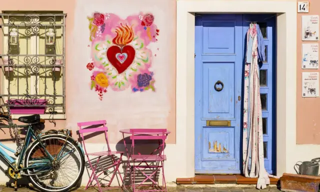 Un angolo pittoresco di Borgo San Giuliano a Rimini: una porta blu, un cuore fiammeggiante dipinto, una bicicletta e sedie rosa.