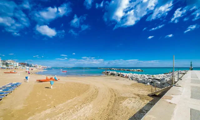 Spiaggia di Bellaria Mare con lettini, bagnini arancioni e mare azzurro sotto un cielo blu con nuvole bianche.