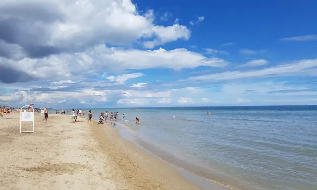 Una giornata di sole a Riccione mare, con gente che passeggia sulla sabbia e si gode l'acqua cristallina sotto un cielo azzurro con nuvole bianche.
