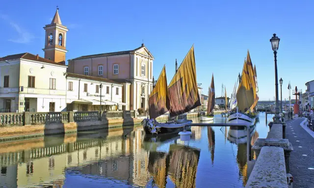 Traditional sailboats with colorful sails line a canal in Cesenatico, Italy, with buildings and a church reflected in the water.
