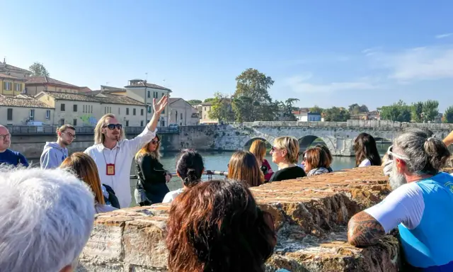 Guida turistica con gruppo a Rimini, che ascolta storie sui **eventi rimini** e la storia locale, con un ponte antico sullo sfondo.