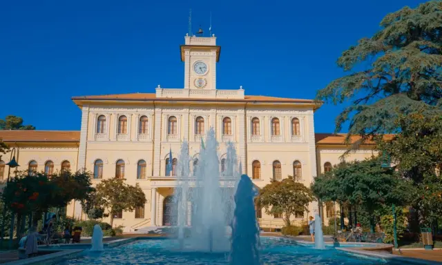 Fontana zampillante nella piazza Roosevelt di Cattolica, con un edificio giallo e un cielo azzurro.