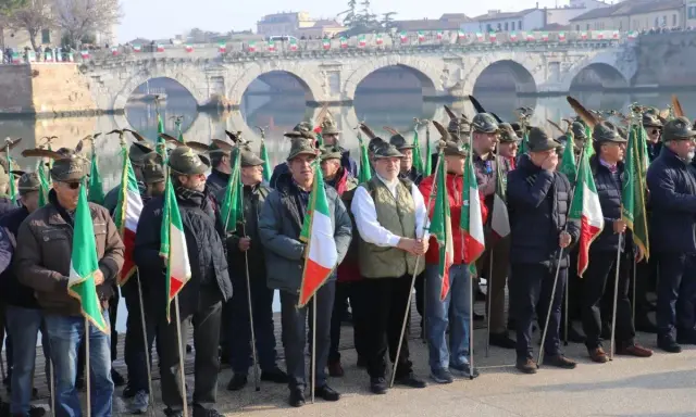 Uomini con cappelli alpini e bandiere italiane partecipano all'adunata alpini a Rimini, con un ponte storico sullo sfondo.