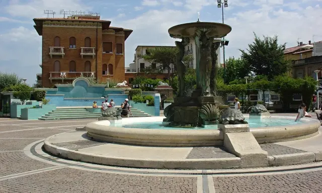 Fontana delle Sirene a Cattolica, con statue di sirene e tartarughe, circondata da edifici storici e una piazza acciottolata.