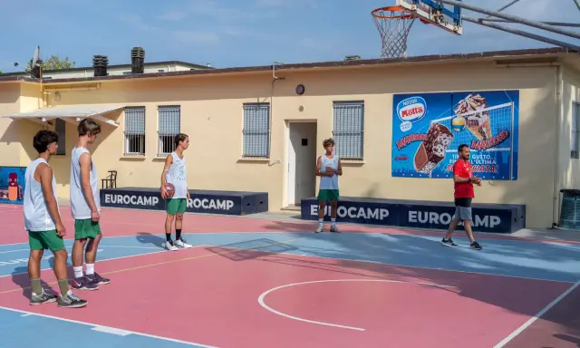Ragazzi in tenuta sportiva giocano a basket in un camp estivo a Cesenatico, con un cartellone pubblicitario di gelati Motta sullo sfondo.