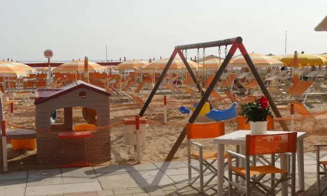 Area giochi e lettini sulla spiaggia 106 di Riccione, con ombrelloni arancioni e bianchi, altalena e casetta per bambini.
